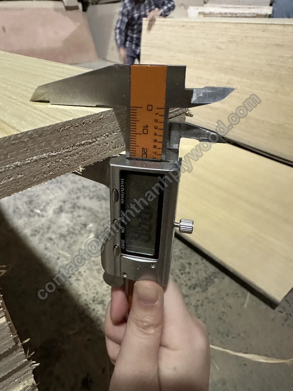 Plywood quality control: digital caliper measuring a finished panel at the edge of a pack on the factory floor